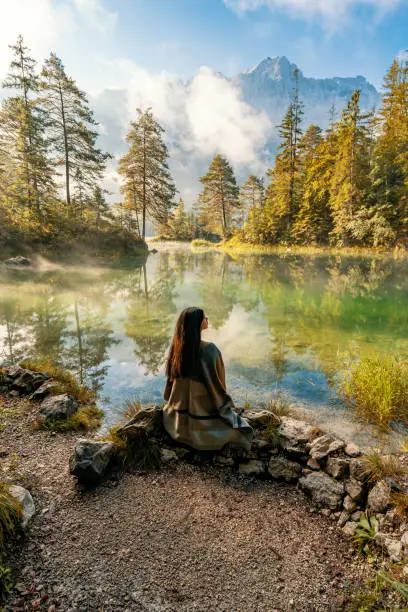 Young woman meditating while sitting, relaxing alone while looking at view, lake and mountains sunny landscape on background outdoor. Travel healthy Lifestyle concept.  Scenic surroundings near famous lake Eibsee. Wonderful day gorgeous scene. Location resort Garmisch-Partenkirchen, Bavarian alp, sightseeing Europe. Outdoor activity. Explore the world's beauty.