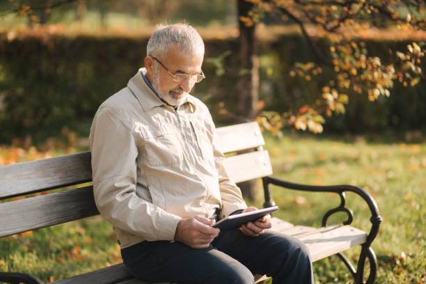 istock Handsome elderly man sitting on the bench and use tablet for scrolling in internet. Background of autumn tree 1184338352