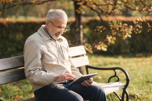 istock Handsome elderly man sitting on the bench and use tablet for scrolling in internet. Background of autumn tree 1184339086