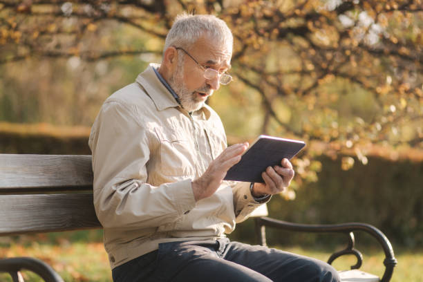 istock Handsome elderly man sitting on the bench and use tablet for scrolling in internet. Background of autumn tree 1184339182