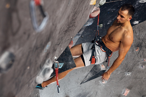 Muscular shitrless man climbing wall with belay device at an indoor wall climbing centre. Rock climber practicing climbing at an indoor climbing gym.