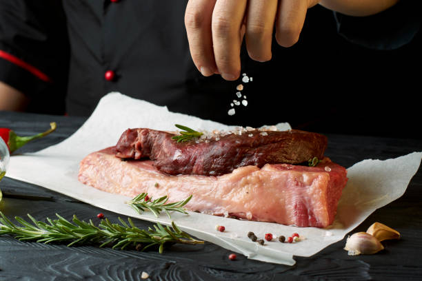 istock Chef salts steak with herbs on paper. Preparing fresh beef or pork. Horizontal photo with a dark black background. 1184361473