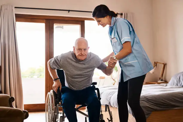 Shot of a young nurse helping a senior man get up from a wheelchair in a retirement home