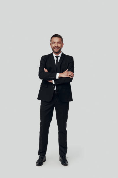 istock Full length of charming young man in full suit smiling and looking at camera while standing against grey background 1184375514