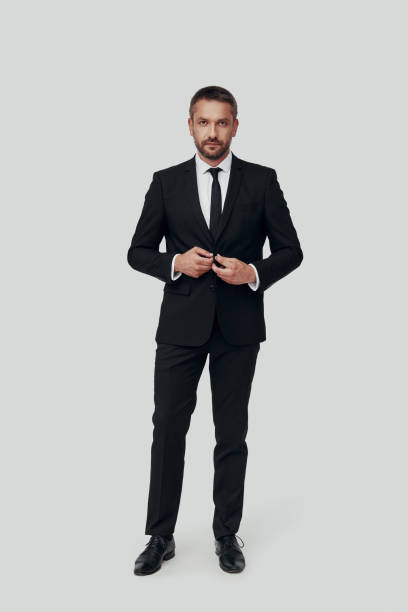 istock Full length of charming young man in full suit adjusting suit and looking at camera while standing against grey background 1184375562
