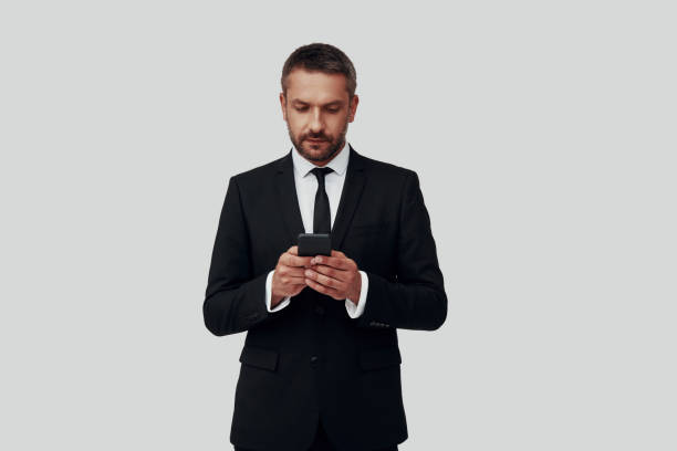 istock Charming young man in full suit using smart phone while standing against grey background 1184378273