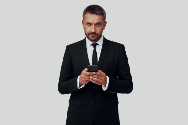 Charming young man in full suit using smart phone while standing against grey background