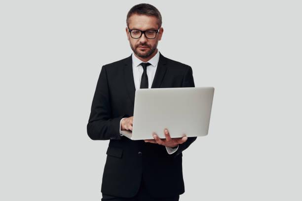 istock Charming young man in full suit using laptop while standing against grey background 1184378672