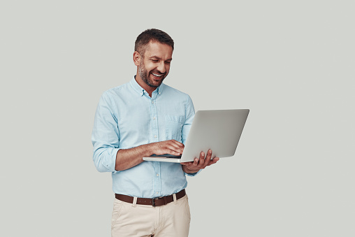 Handsome young man using laptop and smiling while standing against grey background