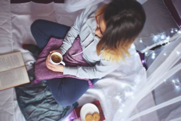 istock Young woman, sitting on a bed holding a coffee mug and reading a book 1184384415