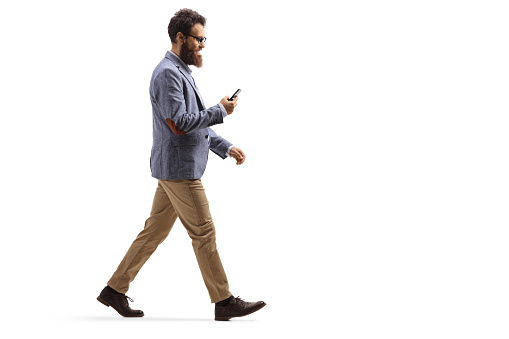 Full length profile shot of a bearded man walking and looking at his mobile phone isolated on white background