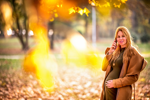 Blond pregnant woman talking on a smart phone while strolling through the park during autumn.