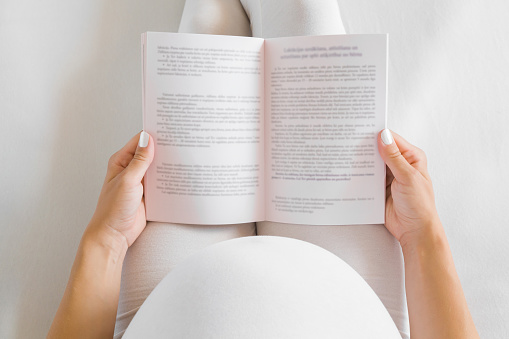 Pregnant woman in white clothes sitting on bed and reading book. Relaxing at home. Baby expectation. Point of view shot. Top view.