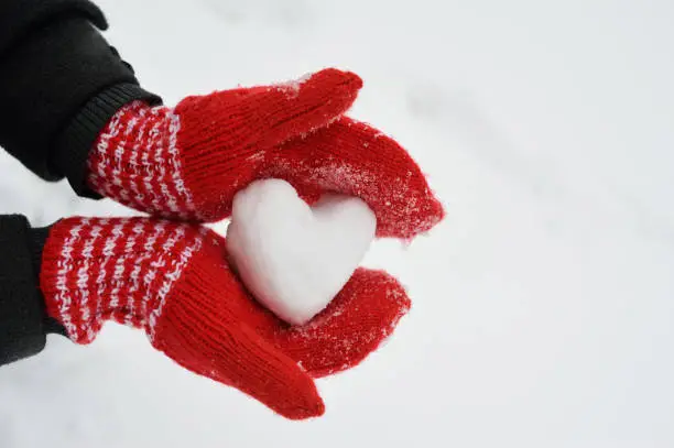 Female hands in warm red crocheted mittens with snowy heart. White snow background. Love concept. Valentine's Day Greeting card with copyspace.