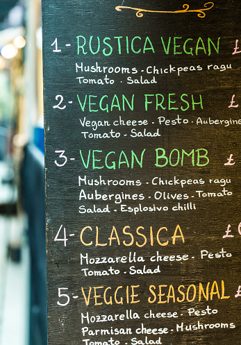 Close up image depicting menu options for a vegan and vegetarian cafe on a city street in central London, UK. Menu options include such things as vegan cheese, mushrooms, aubergines and olives. Focus is on the sign with colorful chalk writing in the foreground, while the street is defocused beyond. Room for copy space.