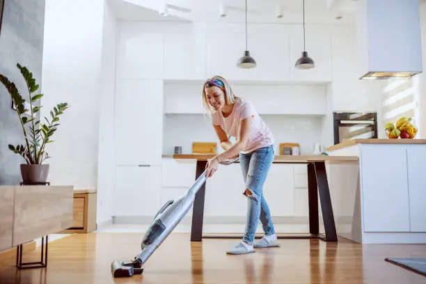 Smiling worthy Caucasian blond housewife dressed casual using steamer to clean parquet in her apartment.