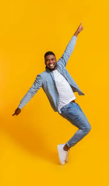 Photo of Cheerful African Man Standing On Tiptoes, Fooling Over Yellow Background