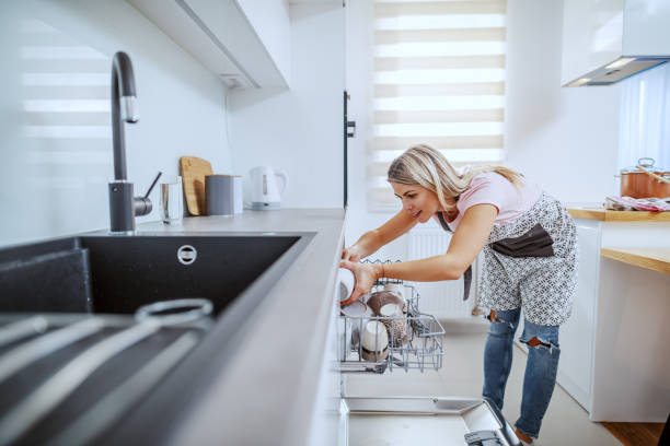 Worthy caucasian blond housewife in apron standing in domestic kitchen and putting dishes in dishwasher. Worthy caucasian blond housewife in apron standing in domestic kitchen and putting dishes in dishwasher. kitchen dishwasher stock pictures, royalty-free photos & images