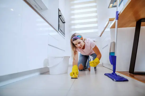 Tidy worthy caucasian blond housewife with rubber gloves kneeling in kitchen and cleaning kitchen floor with sponge and sprayer.