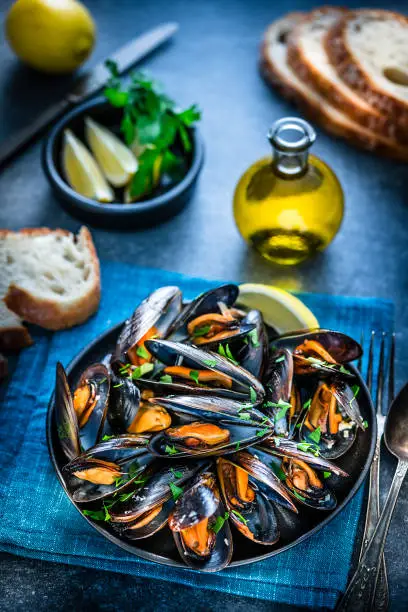 High angle view of steamed mussels in a black plate shot on bluish tint background. Sliced lemon and bread pieces complete the composition. Selective focus on mussels. Predominant colors are black, orange and blue. XXXL 42Mp studio photo taken with Sony A7rii and Sony FE 90mm f2.8 macro G OSS lens