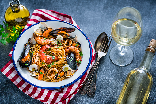 Typical Italian food: seafood pasta plate shot from above on gray abstract background. A white wineglass and wine bottle are beside the plate. The plate is at the left placed on a red and white checkered tablecloth. An olive oil bottle and a parsley twig complete the composition. XXXL 42Mp studio photo taken with Sony A7rii and Sony FE 90mm f2.8 macro G OSS lens