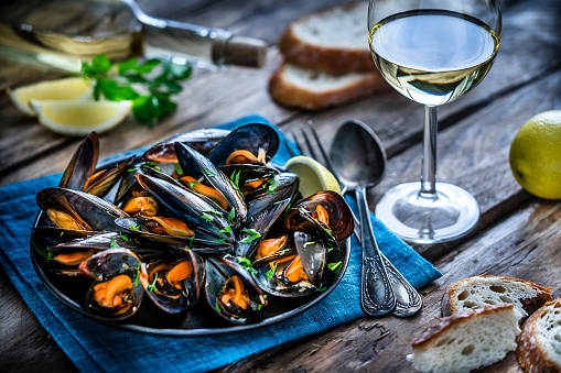 High angle view of steamed mussels in a black plate and white wine glass shot on rustic wooden table. Sliced lemon and bread pieces complete the composition. Selective focus on mussels. Predominant colors are black, orange and blue. XXXL 42Mp studio photo taken with Sony A7rii and Sony FE 90mm f2.8 macro G OSS lens