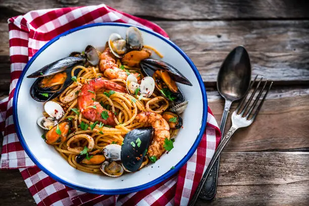 Typical Italian food: seafood pasta plate shot from above on rustic wooden table.  The plate is at the left placed on a red and white checkered tablecloth. XXXL 42Mp studio photo taken with Sony A7rii and Sony FE 90mm f2.8 macro G OSS lens