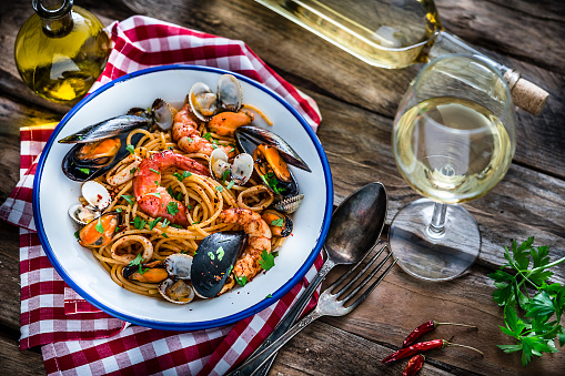 Typical Italian food: seafood pasta plate shot from above on rustic wooden table. A white wineglass and wine bottle are beside the plate. The plate is at the left placed on a red and white checkered tablecloth. An olive oil bottle, parsley twig and red chili peppers complete the composition. XXXL 42Mp studio photo taken with Sony A7rii and Sony FE 90mm f2.8 macro G OSS lens