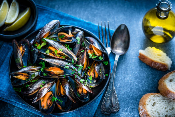 High angle view of steamed mussels in a black plate shot on bluish tint background. Sliced lemon and bread pieces complete the composition. Selective focus on mussels. Predominant colors are black, orange and blue. XXXL 42Mp studio photo taken with Sony A7rii and Sony FE 90mm f2.8 macro G OSS lens