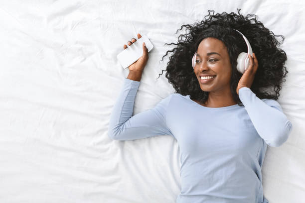 happy african girl listening to music on smartphone in bed - young adult technology beautiful singing imagens e fotografias de stock