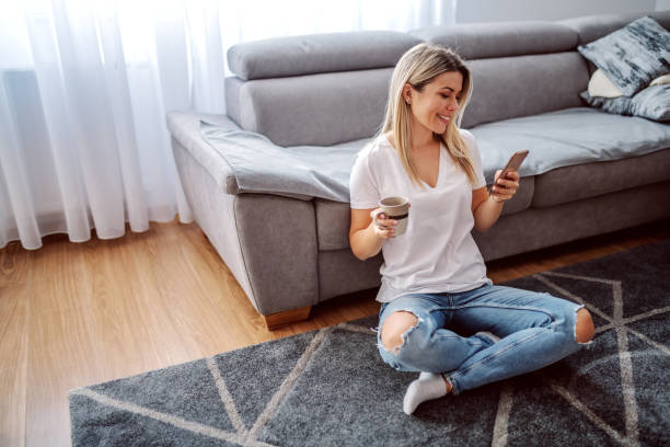 encantadora mujer rubia sonriente caucásica vestida casual sentada en el suelo en la sala de estar con las piernas cruzadas, bebiendo café y usando el teléfono inteligente. - young women sitting simple living eastern europe fotografías e imágenes de stock