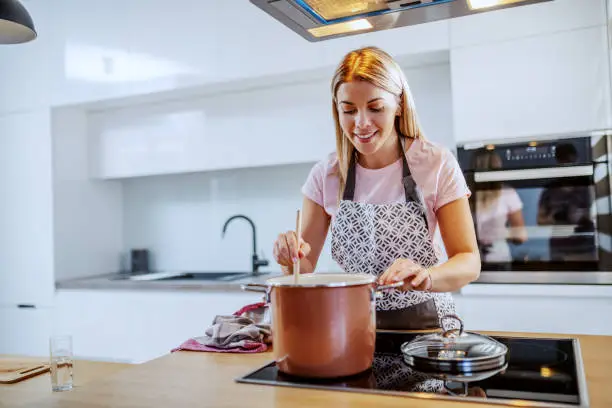 Beautiful smiling caucasian blond young woman in apron standing in kitchen and stirring soup in pot.