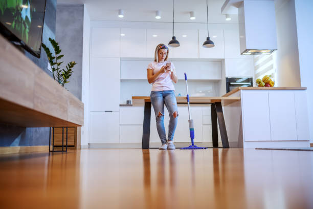 istock Beautiful Caucasian blond housewife dressed casual leaning on dining table and using smart phone for sending message. Next to her is mop. 1184414362
