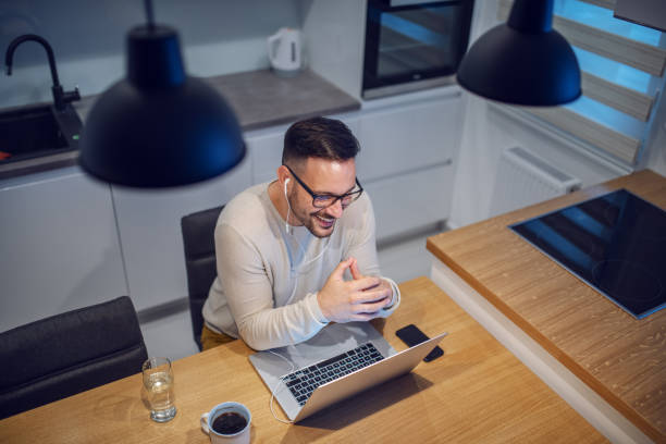 vue supérieure de l'homme caucasien de sourire attrayant s'asseyant à la table de salle à manger dans la cuisine et ayant l'appel vidéo avec sa amie. sur la table sont ordinateur portable, verre avec de l'eau et tasse avec du café. - hot drink audio photos et images de collection