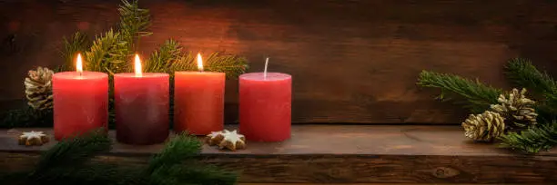 Third Advent, three of four red candles are lit with a flame, fir branches and Christmas decoration on dark rustic wood, wide panoramic format with copy space, selected focus