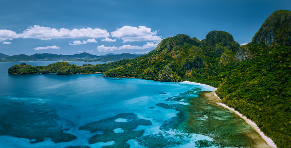 Aerial drone panoramic view of uninhabited tropical island with rugged mountains, rainforest jungle and big blue bay with shallow ocean water.