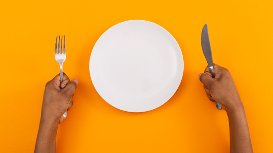 Hunger concept. Black woman impatiently waiting for food with empty plate and holding fork and knife in hands over orange background, top view