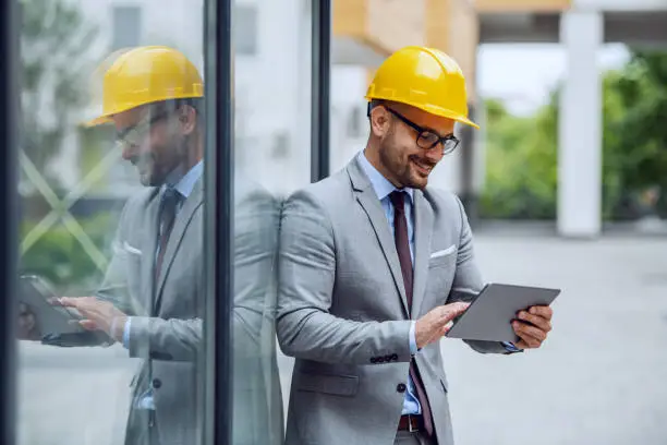 Photo of Handsome caucasian smiling elegant architect in suit, with eyeglasses and helmet on head leaning on window at construction site and using tablet.