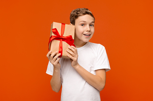 Curious teen boy checking gift, shaking present box, orange background