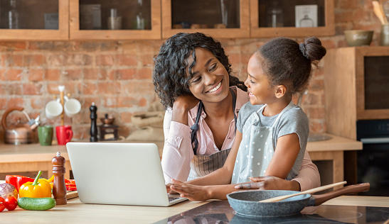 Black woman and her cute daughter embracing and reading recipe on laptop, cooking at kitchen, copy space