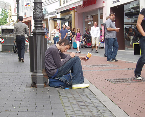 Bonn, Nordrhein Westfalen, Germany, 08/06/2012: Homeless young man asks for alms on the street. A homeless sad man with his head bowed sits on his backpack near a post on the street, in a pedestrian zone with an outstretched hand. In the hand is a coffee cup. People walk around.
