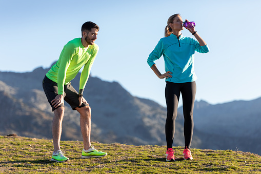 Shot of healthy young couple relaxing after running while the woman drinks water on the mountain.
