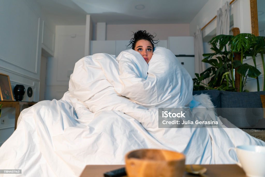 Young attractive anxious woman watching TV series and wrapping in big white blanket. cold autumn or winter time spending Young attractive anxious woman watching TV series and wrapping in big white blanket. Concept of cold autumn or winter time spending. Modern interior Cold Temperature Stock Photo