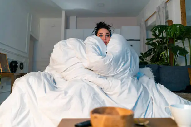 Photo of Young attractive anxious woman watching TV series and wrapping in big white blanket. cold autumn or winter time spending