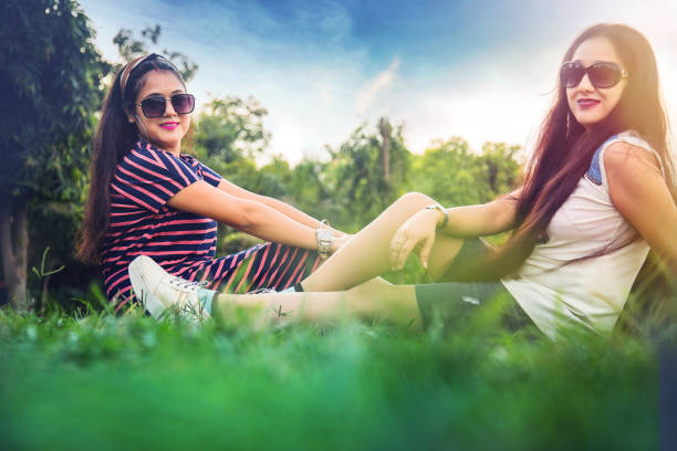 Two happy pretty women posing in park. Women sitting  on green grass and looking at camera. Women friendship and nature concept. side view.