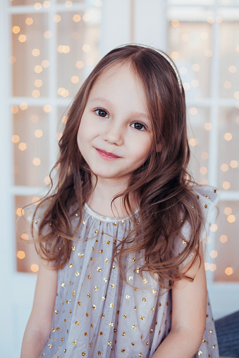 Portrait of cute smiling child girl wearing sparkling dress posing against christmas background. Xmas holiday indoor photoshot. Vertical image in soft tones.