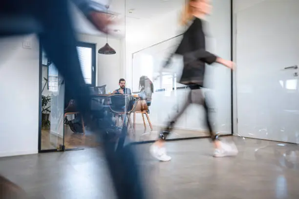 Blurred motion of colleagues walking briskly down office hallway as colleagues sit in conference room discussing ideas.