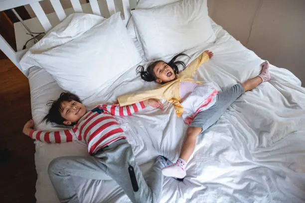 High angle view of excited young Chinese brother and sister laying spreadeagled on messy guest bed.