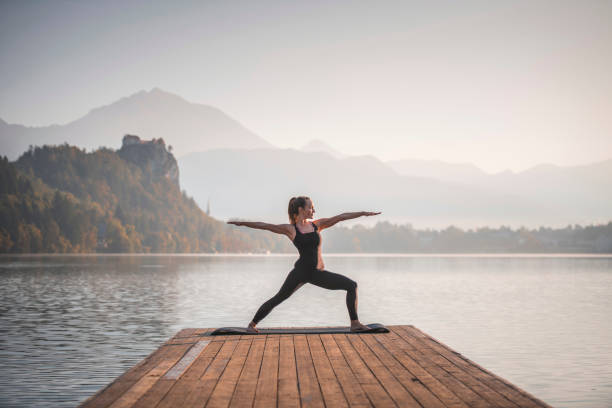 młoda kobieta w warrior ii pozycja z widokiem na jezioro bled - posture women side view yoga zdjęcia i obrazy z banku zdjęć