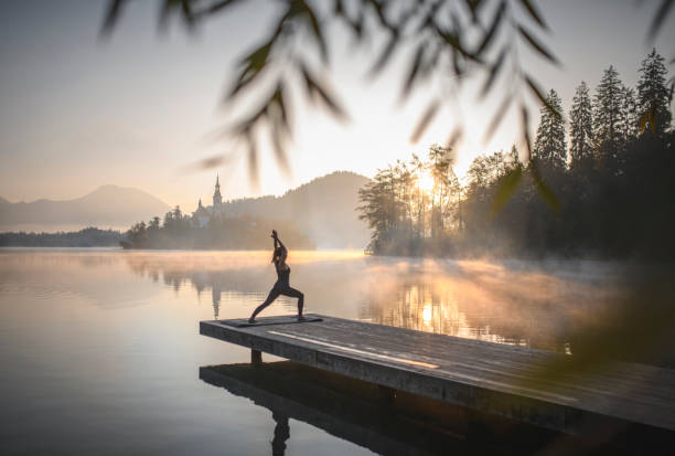 Young Woman in Yoga High Lunge Position at Dawn in Bled Mid distance view of young Caucasian woman standing on pier overlooking misty Lake Bled in yoga High Lunge Position (Utthita Ashwa Sanchalanasana) at dawn. gorenjska stock pictures, royalty-free photos & images